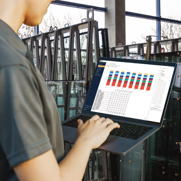 Person arbeitet an einem Laptop mit Datenanalyse-Dashboard in einer industriellen Umgebung mit Glasscheiben im Hintergrund