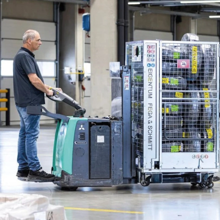 box-id_success-story-fega-schmitt_warehouse-worker-pallet-truck Lagerarbeiter transportiert Gitter-Ladungsträger mit Niederhubwagen in Halle
